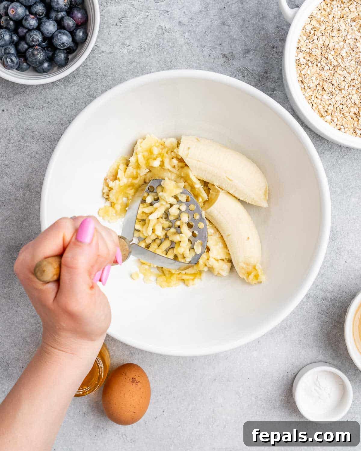 Soft-Baked Blueberry Oatmeal Cookies 4 A close-up view of a banana being mashed in a mixing bowl with a potato masher, illustrating the first step of the recipe.