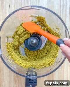 Spatula scraping down the blended pistachio in the food processor.