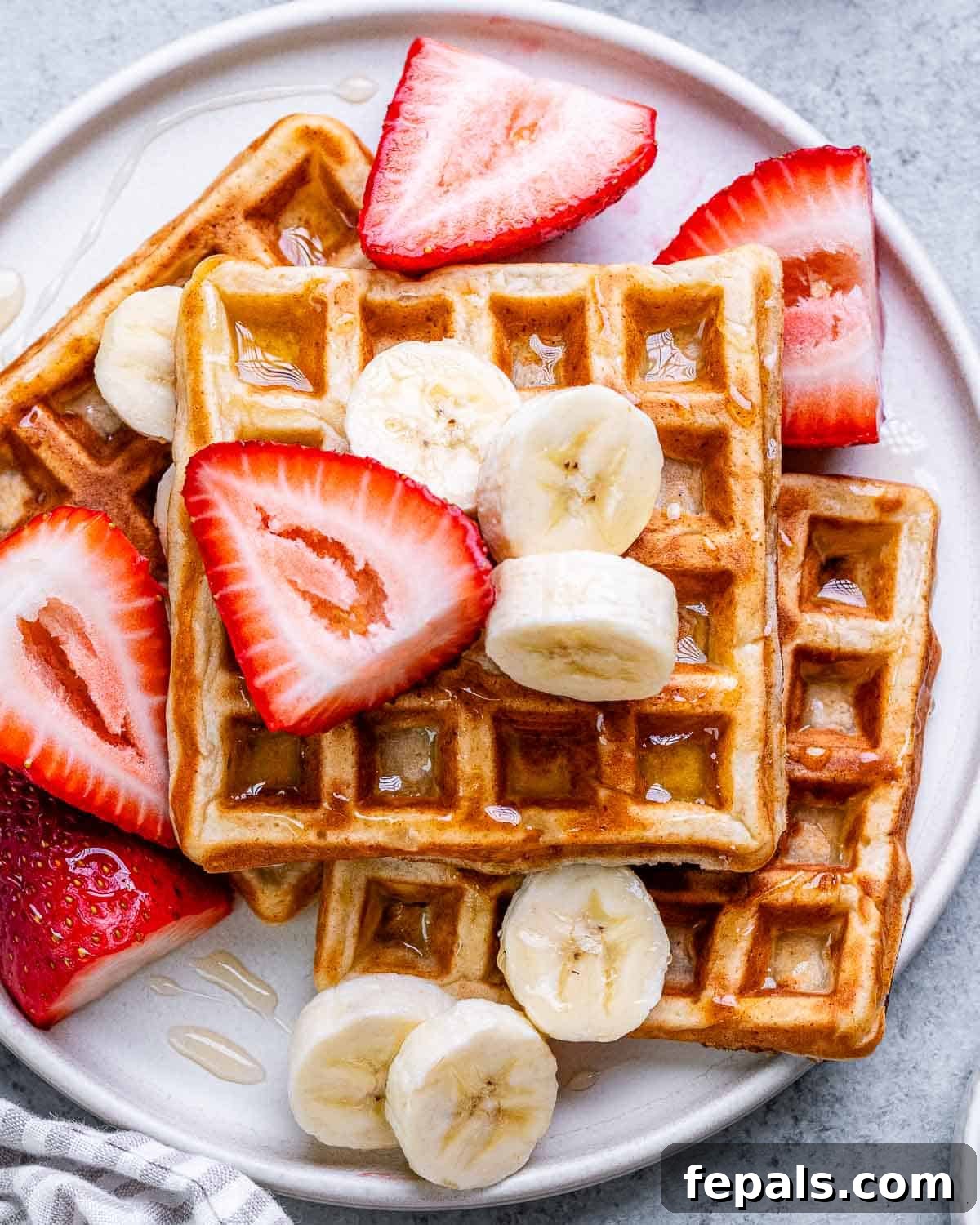 Golden-brown cottage cheese waffles on a white plate, generously topped with sliced fresh strawberries and ripe bananas, with a drizzle of syrup.