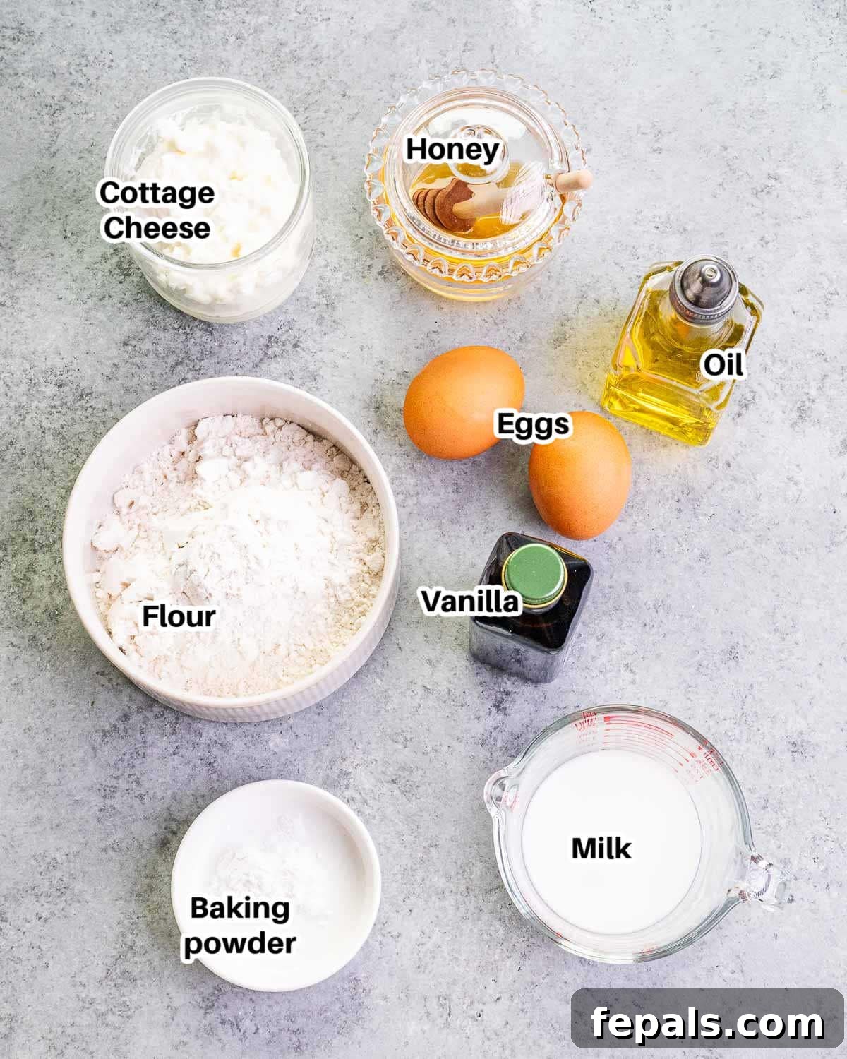 A collection of ingredients laid out on a countertop, including a bowl of cottage cheese, eggs, vanilla extract, honey, milk, vegetable oil, flour, and baking powder, ready to make waffles.