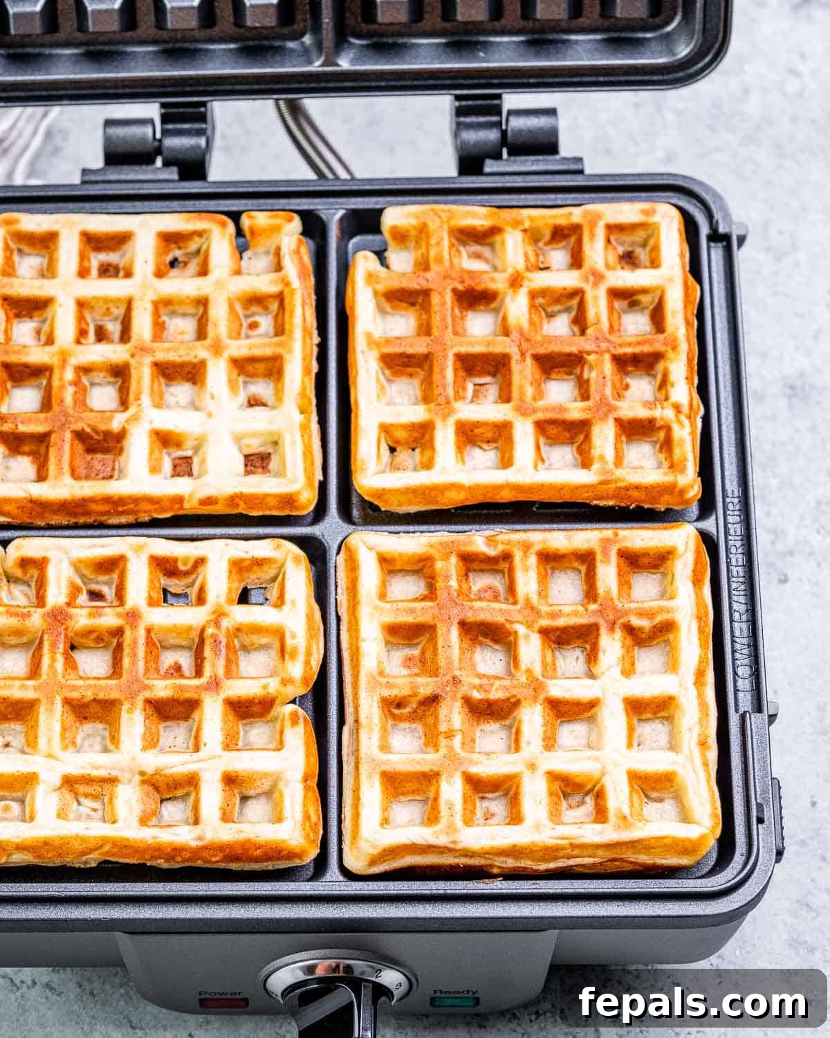 Freshly cooked, golden-brown waffles are shown on an open waffle maker, steam gently rising.