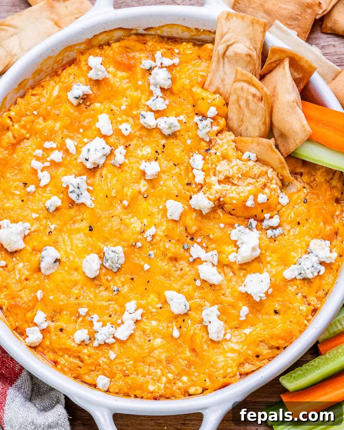 Close-up of the baked cottage cheese buffalo chicken dip, showing the melted cheddar and crumbled blue cheese topping, with pita chips and celery sticks blurred in the background.