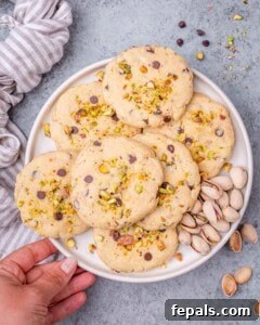 A hand holding a plate of baked pistachio chocolate chip cookies, with a linen napkin in the background and shelled pistachios nearby.
