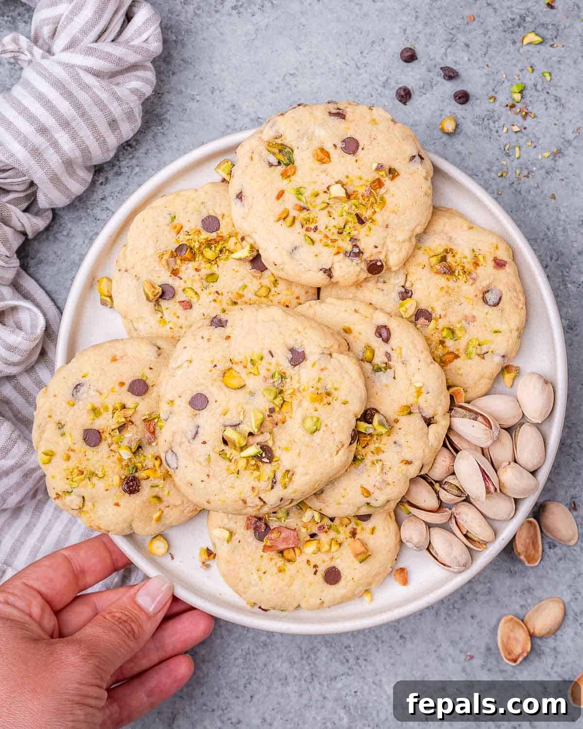 Decadent Pistachio Chocolate Chip Cookies 5 A hand holding a plate of baked pistachio chocolate chip cookies, with a linen napkin in the background and shelled pistachios nearby.