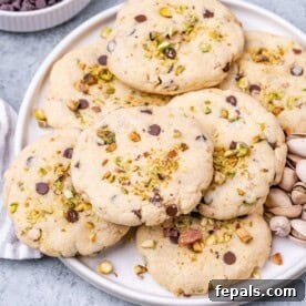 A white plate filled with pistachio chocolate chip cookies, topped with chopped pistachios and surrounded by shelled pistachios.