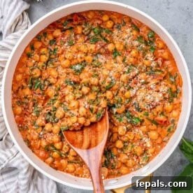 Wooden spoon stirring creamy chickpeas and spinach in a pan with a tomato-based sauce.