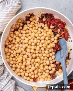 Chickpeas and chopped sun dried tomatoes being stirred together in a white skillet.