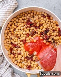 Chickpeas and sundried tomatoes in a pan with tomato passata being added to the skillet before mixing.