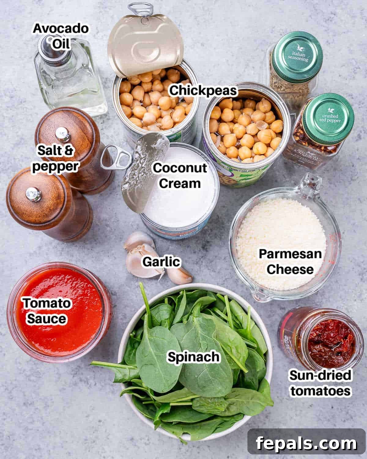A vibrant display of fresh ingredients for Marry Me Chickpeas, including avocado oil, garlic, sun-dried tomatoes, Italian seasoning, chili flakes, canned chickpeas, tomato sauce, coconut cream, baby spinach, Parmesan, and fresh basil.