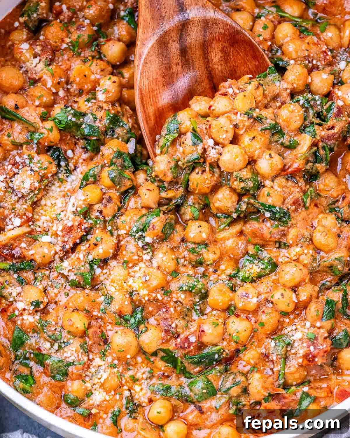 A wooden spoon scooping a generous portion of creamy Marry Me Chickpeas, featuring visible spinach and melted Parmesan, from a rustic skillet.