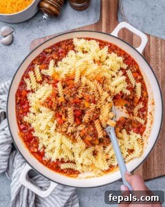 Cooked pasta being folded into the skillet with the sauce and meat.