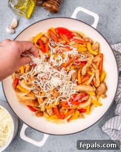 Shredded mozzarella cheese being sprinkled by hand over a mix of cooked chicken and raw peppers in a white bowl.