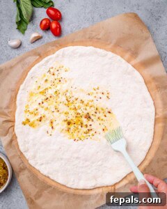 Raw pizza dough spread out on parchment paper with garlic oil being brushed over the top.