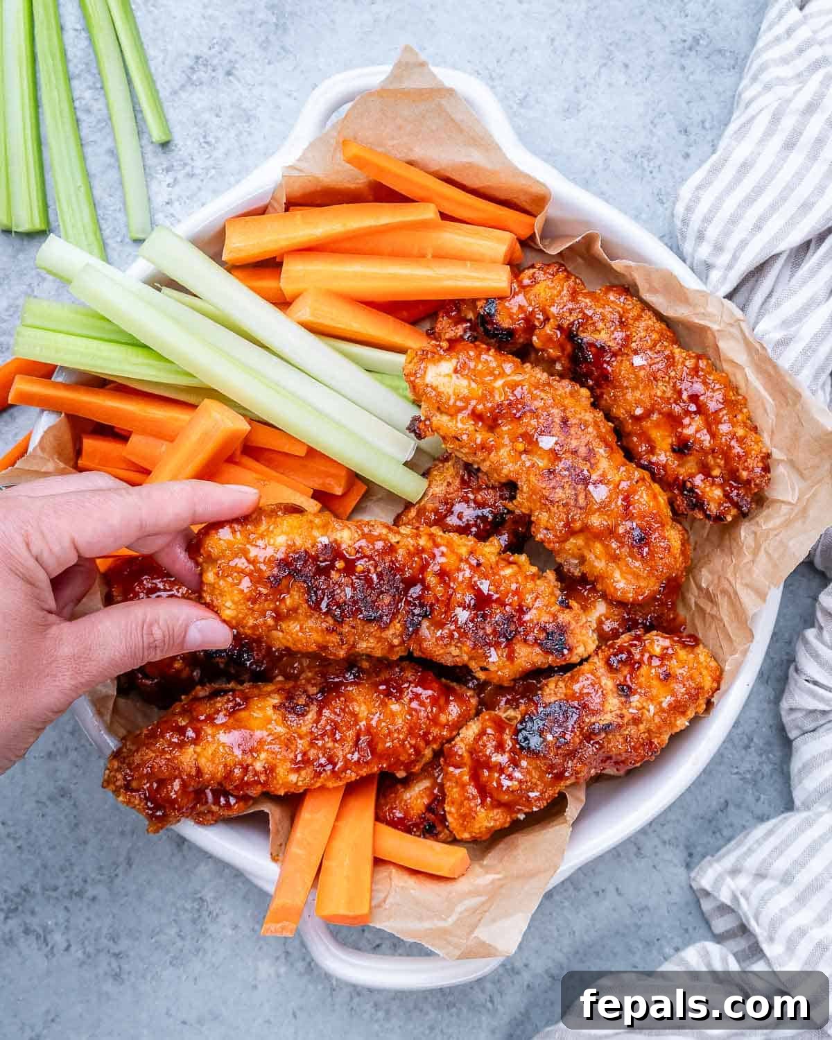 Zesty Air Fried Buffalo Chicken Strips 2 Fully cooked buffalo chicken tenders served in a bowl with fresh celery and carrot sticks, hand reaching in to grab one.