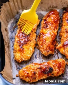 Crispy buffalo chicken tenders on parchment paper being brushed with buffalo sauce using a yellow silicone brush.