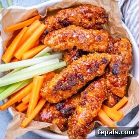 Pile of buffalo chicken tenders on parchment paper, served with carrot and celery sticks in a rustic basket.
