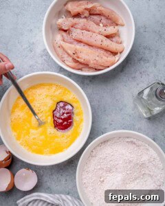 Chicken tenders in a bowl being seasoned with salt and pepper next to bowls of beaten egg and flour mixture.