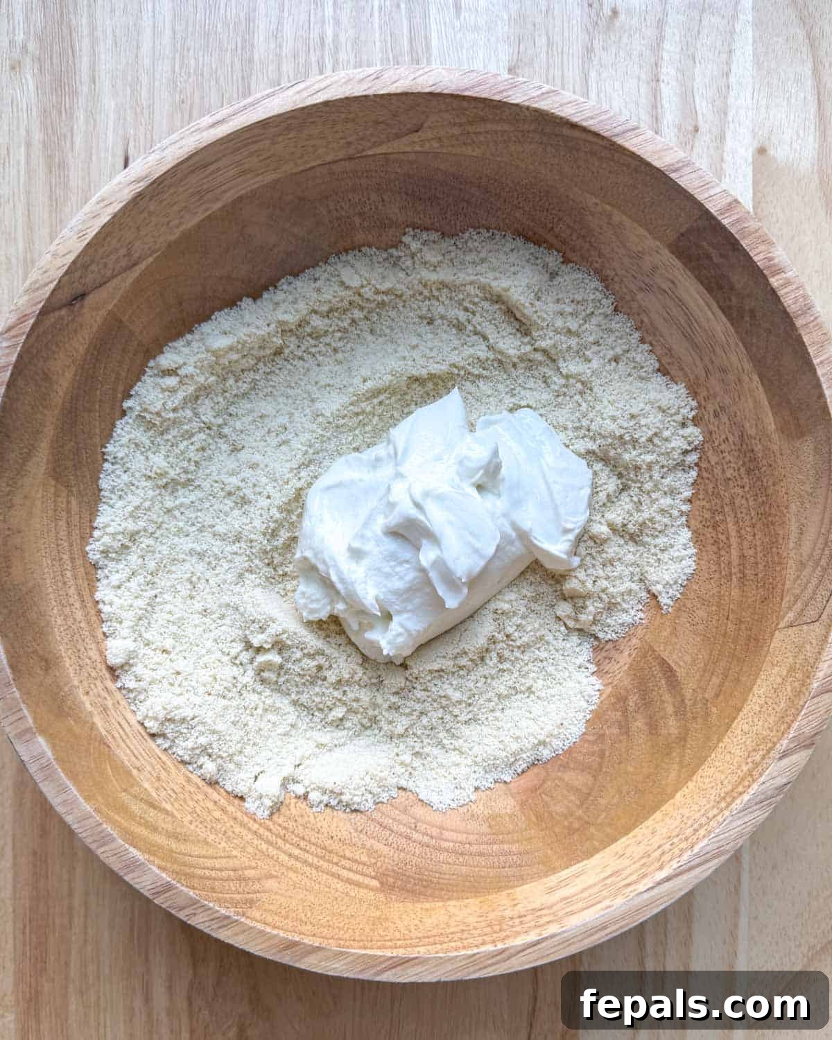 Low-Carb Almond Bagels 6 Greek yogurt being added to the almond flour mixture in a wooden bowl.