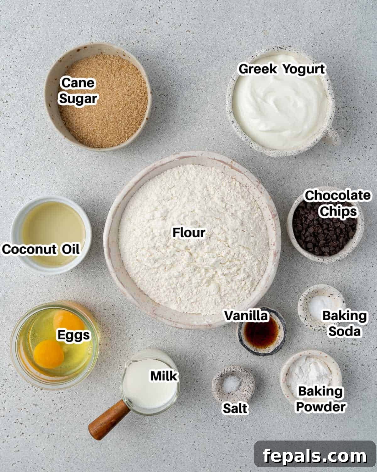 Ingredients for Greek yogurt chocolate chip muffins, including flour, sugar, baking agents, oil, yogurt, milk, eggs, vanilla, and chocolate chips, arranged on a kitchen counter.