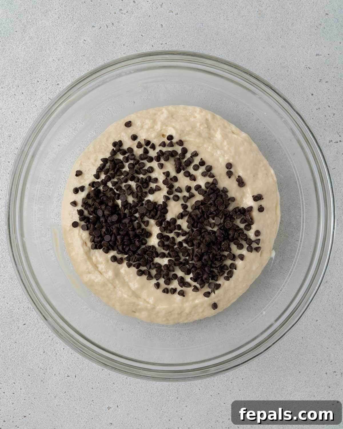 Glass bowl filled with muffin batter topped with mini chocolate chips, ready to be folded in