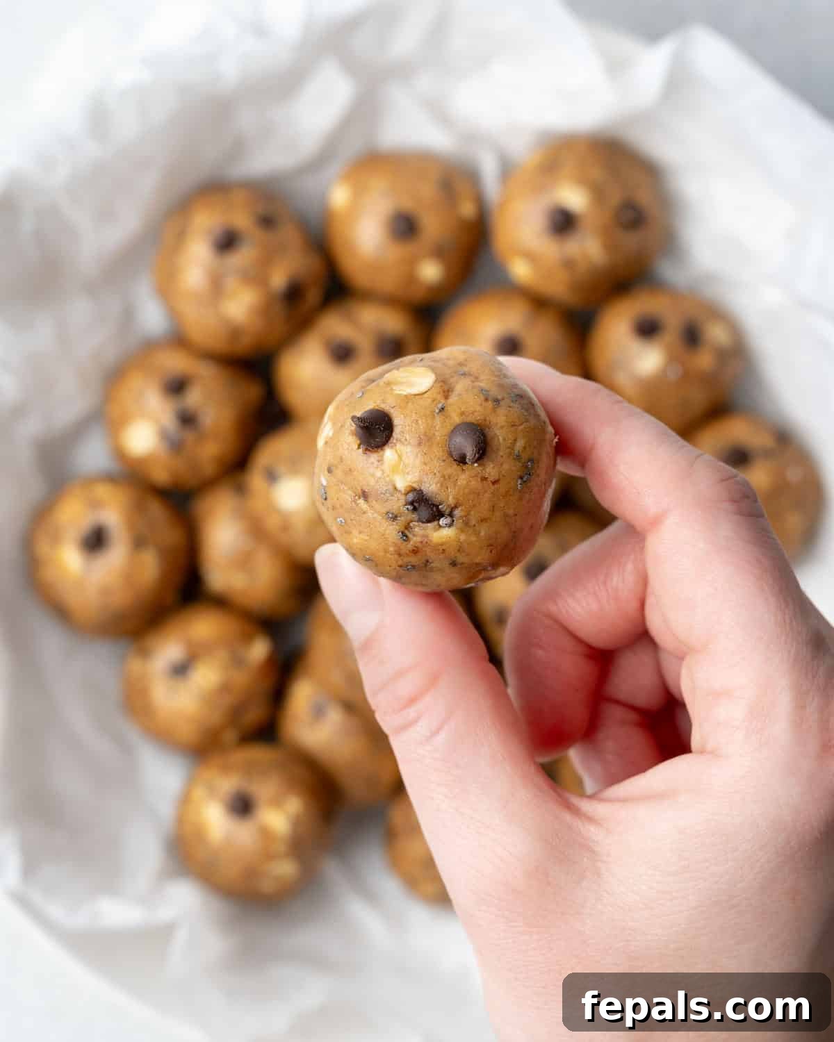 A hand holding one perfectly formed peanut butter protein ball over a blurred bowl.