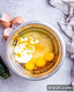 Moist Zucchini Carrot Loaf 8 Overhead view of a mixing bowl filled with eggs, Greek yogurt, vegetable oil, and vanilla extract before whisking, with eggshells and a zucchini nearby on a light kitchen surface.