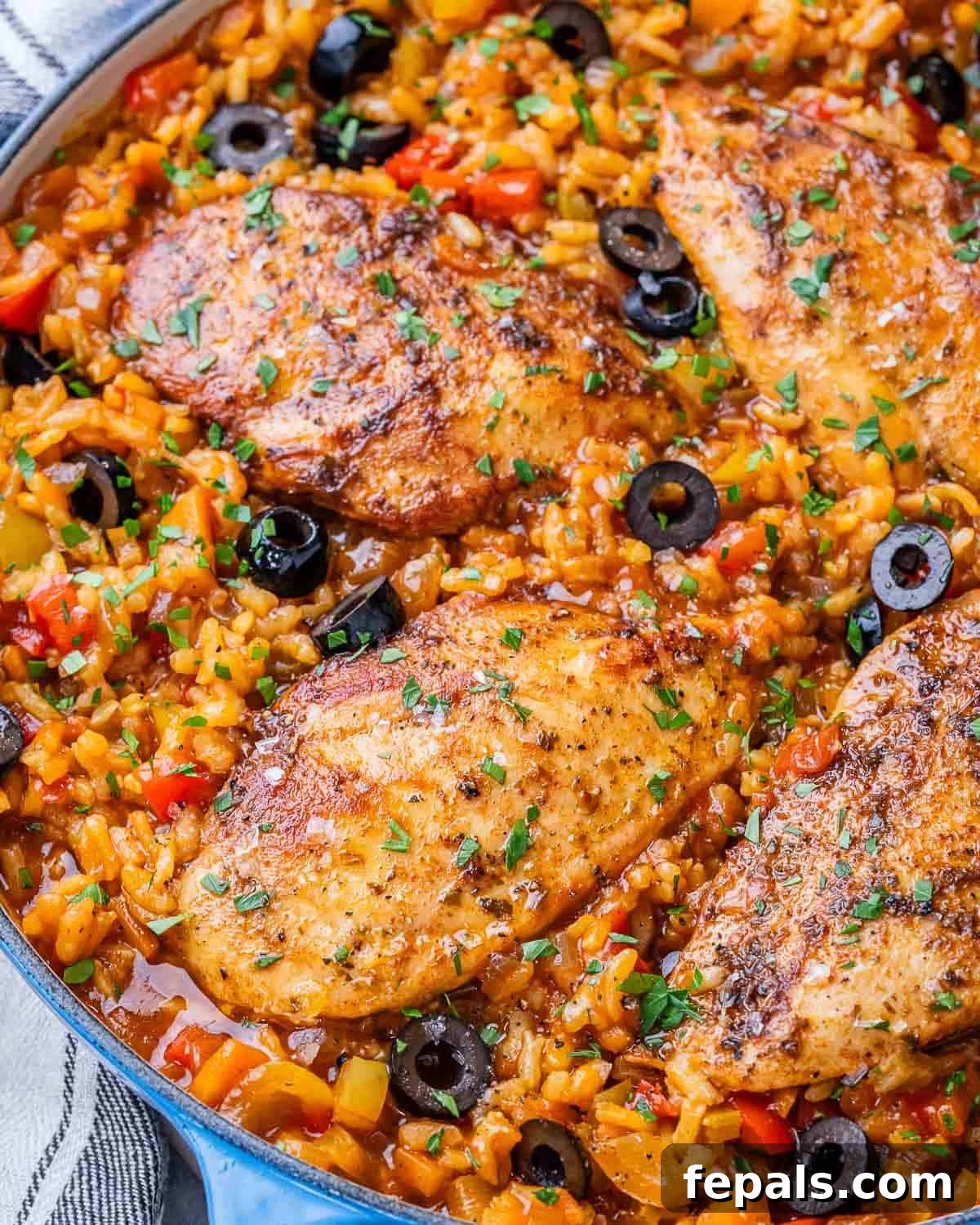 Close-up of golden, seasoned chicken breasts nestled in Spanish rice with black olives and chopped parsley, ready to be served from a skillet.