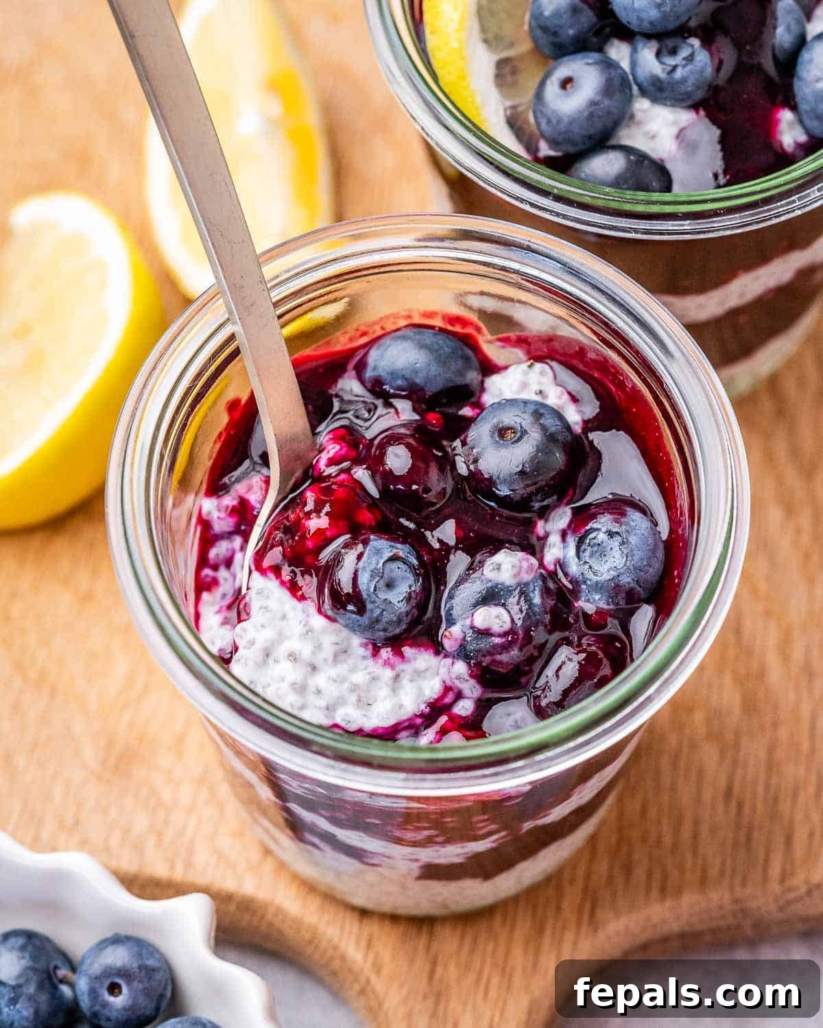 Spoon scooping blueberries from a chia pudding jar, with lemon slices and fresh blueberries in the background on a wooden board.
