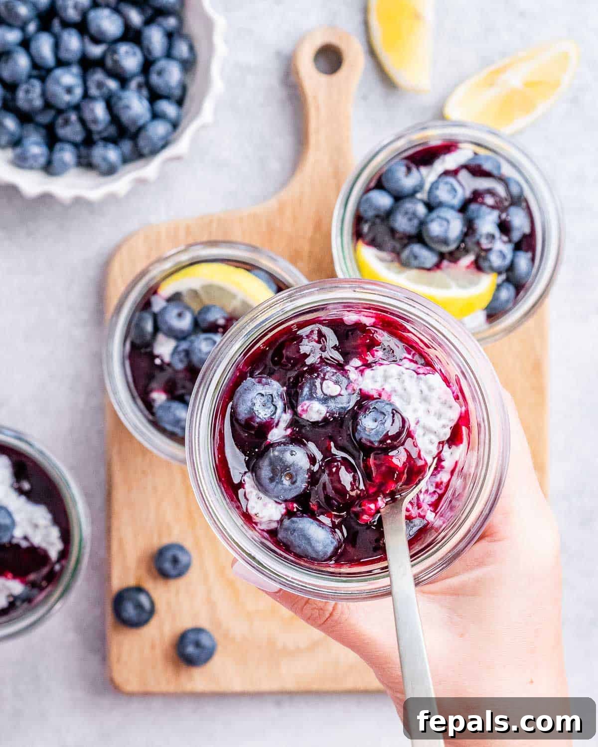 Overhead view of a hand spooning blueberry compote into a glass jar filled with chia pudding, surrounded by fresh ingredients.