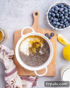 Hand pouring milk into the chia pudding mixture in a white bowl, surrounded by blueberries, honey, and lemons.