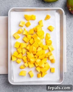 Cubed mangoes spread on a parchment-lined baking sheet, ready for freezing.