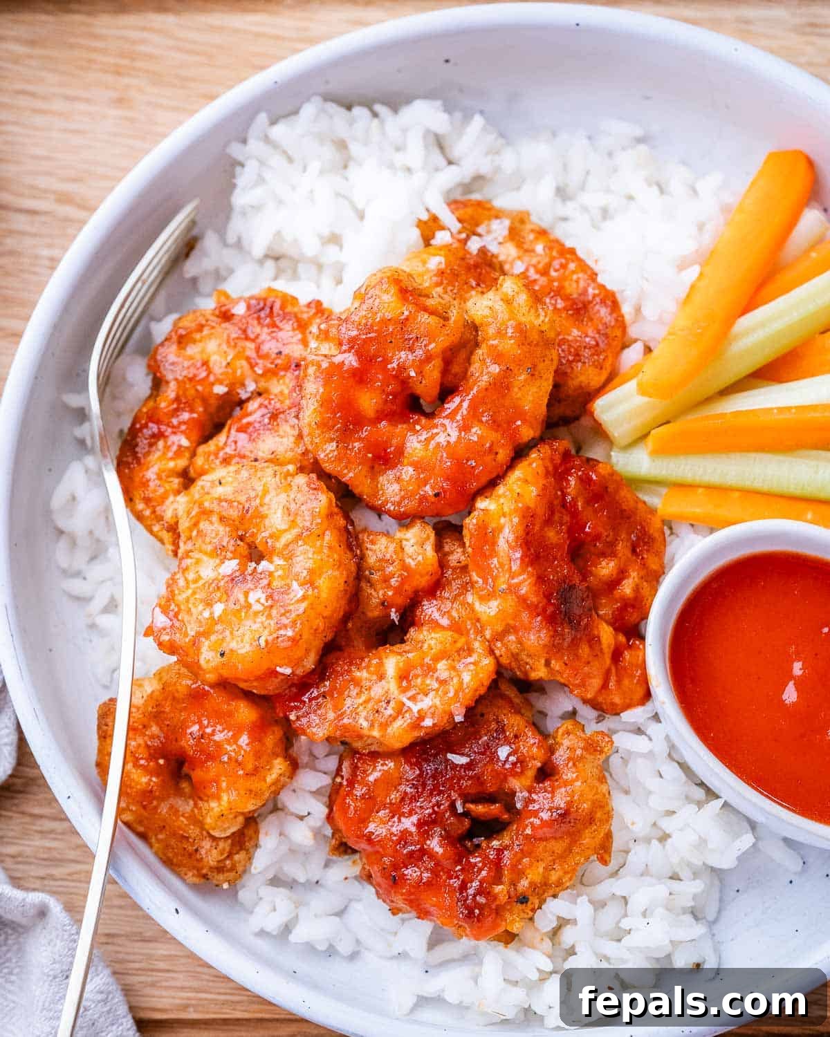 A beautifully arranged bowl of buffalo shrimp served over rice, with fresh celery, carrots, and a small side of extra buffalo sauce.