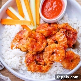 A bowl of rice topped with buffalo shrimp, accompanied by celery, carrots, and a small bowl of extra buffalo sauce.