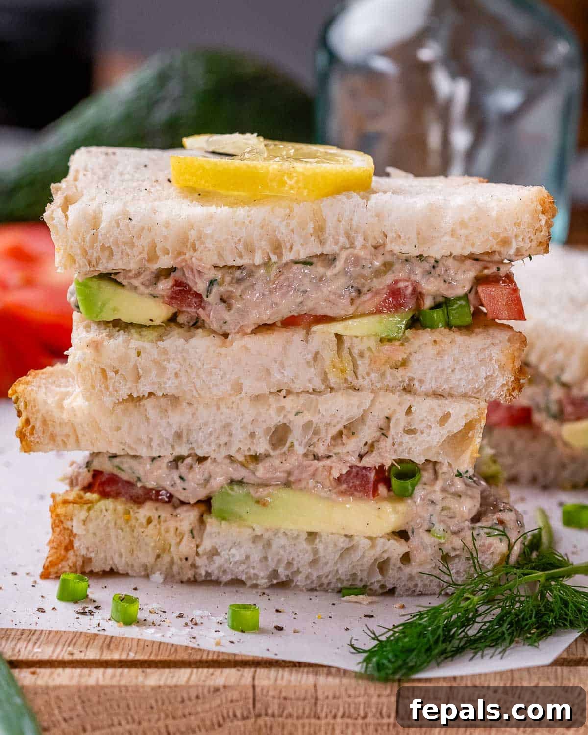 Two stacked halves of a tunacado sandwich on toasted bread, garnished with a lemon slice, fresh dill, and green onion.