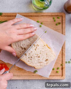 Overhead shot of a hand slicing the sandwich diagonally on parchment paper set on a wooden cutting board.