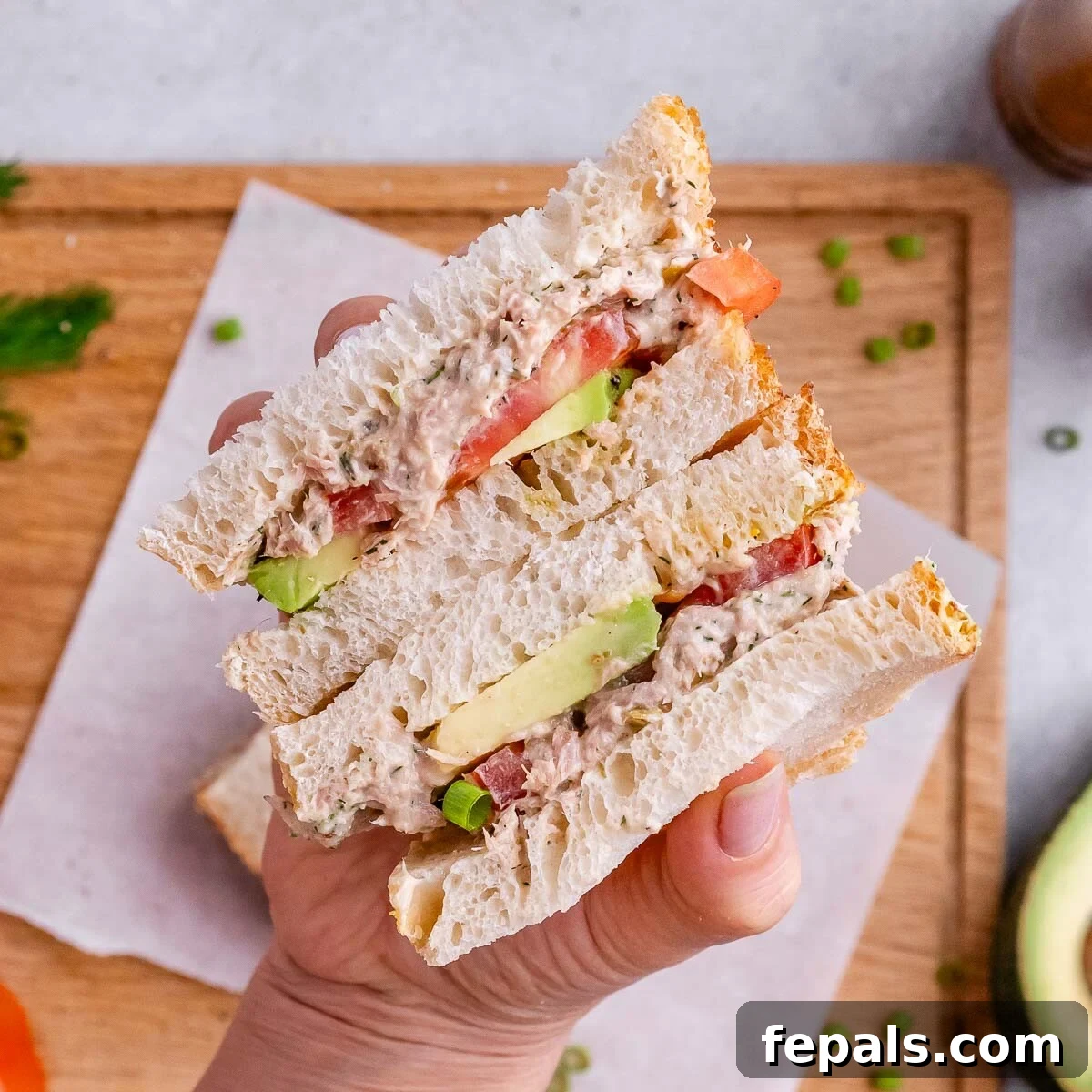 A hand holding a cut sandwich with avocado and tomato layers, placed on parchment over a wooden board.
