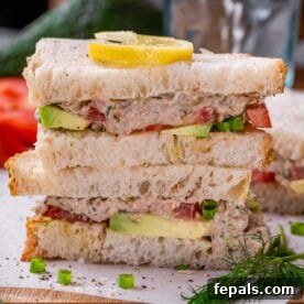 Two stacked halves of a tunacado sandwich on toasted bread, garnished with a lemon slice, fresh dill, and green onion.