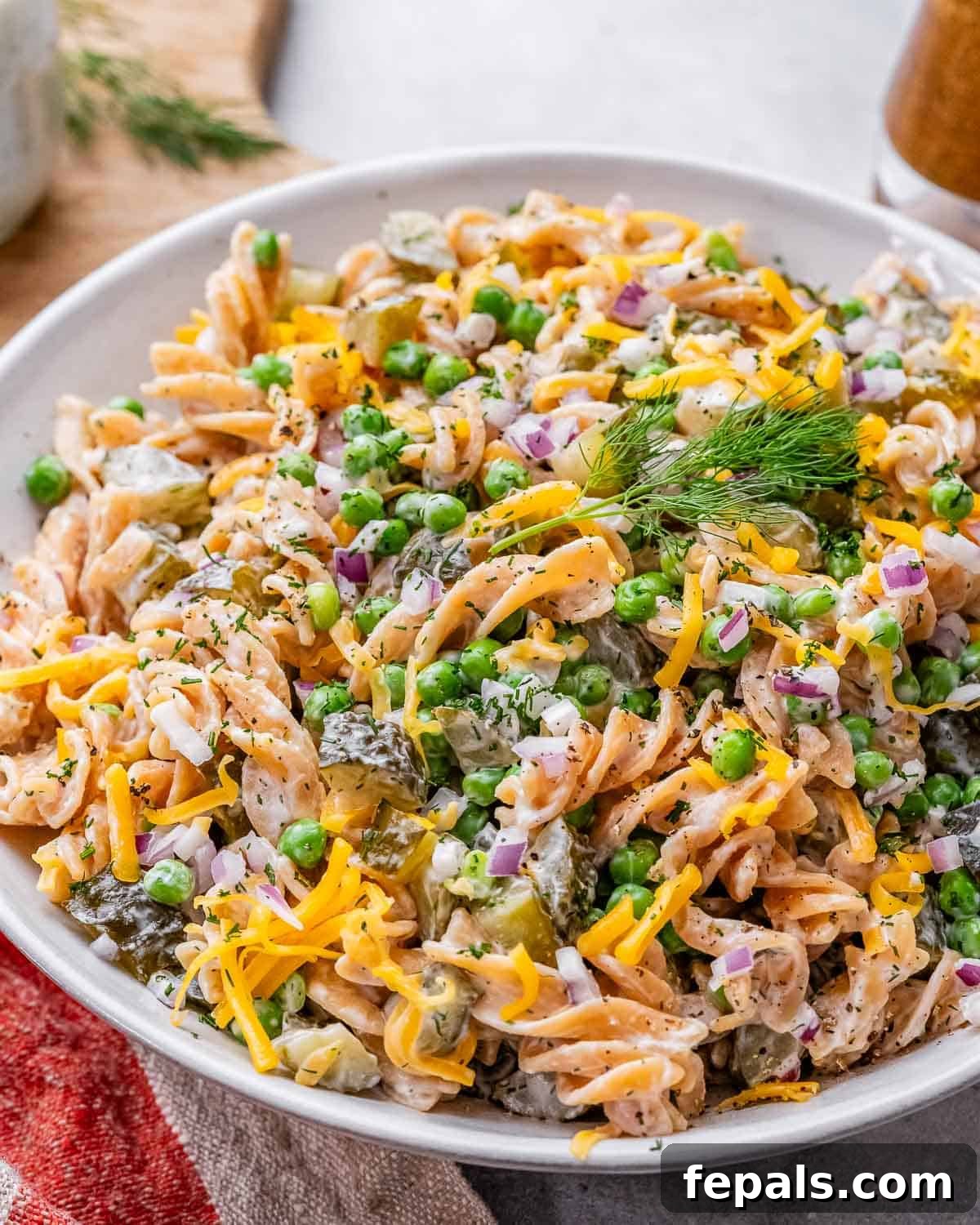 Bowl of finished dill pickle pasta salad, garnished with extra dill and peas, with a spoon ready to serve.