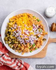 Pasta salad base in a white bowl topped with shredded cheddar, chopped red onion, and diced dill pickles before mixing.