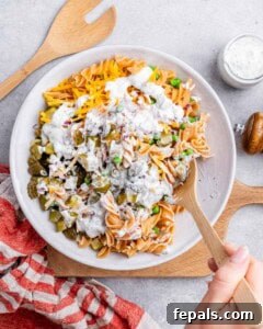 Hand stirring in creamy dressing to the pasta salad mixture in a large white bowl on a wooden board.