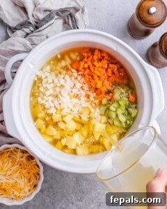 The full crockpot filled with potatoes, chopped carrots, celery, onion, with broth being poured in.