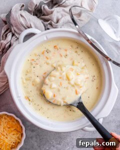Crockpot filled with cooked diced potatoes, carrots, and broth, with a ladle lifting a creamy portion.