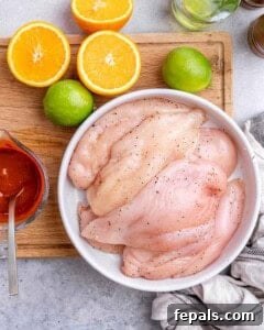 Plain raw chicken breasts in a white bowl, placed next to halved oranges and limes on a wooden board.