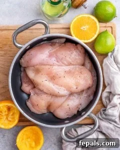 Raw chicken breasts seasoned with salt and pepper in a bowl, surrounded by fresh oranges and limes.