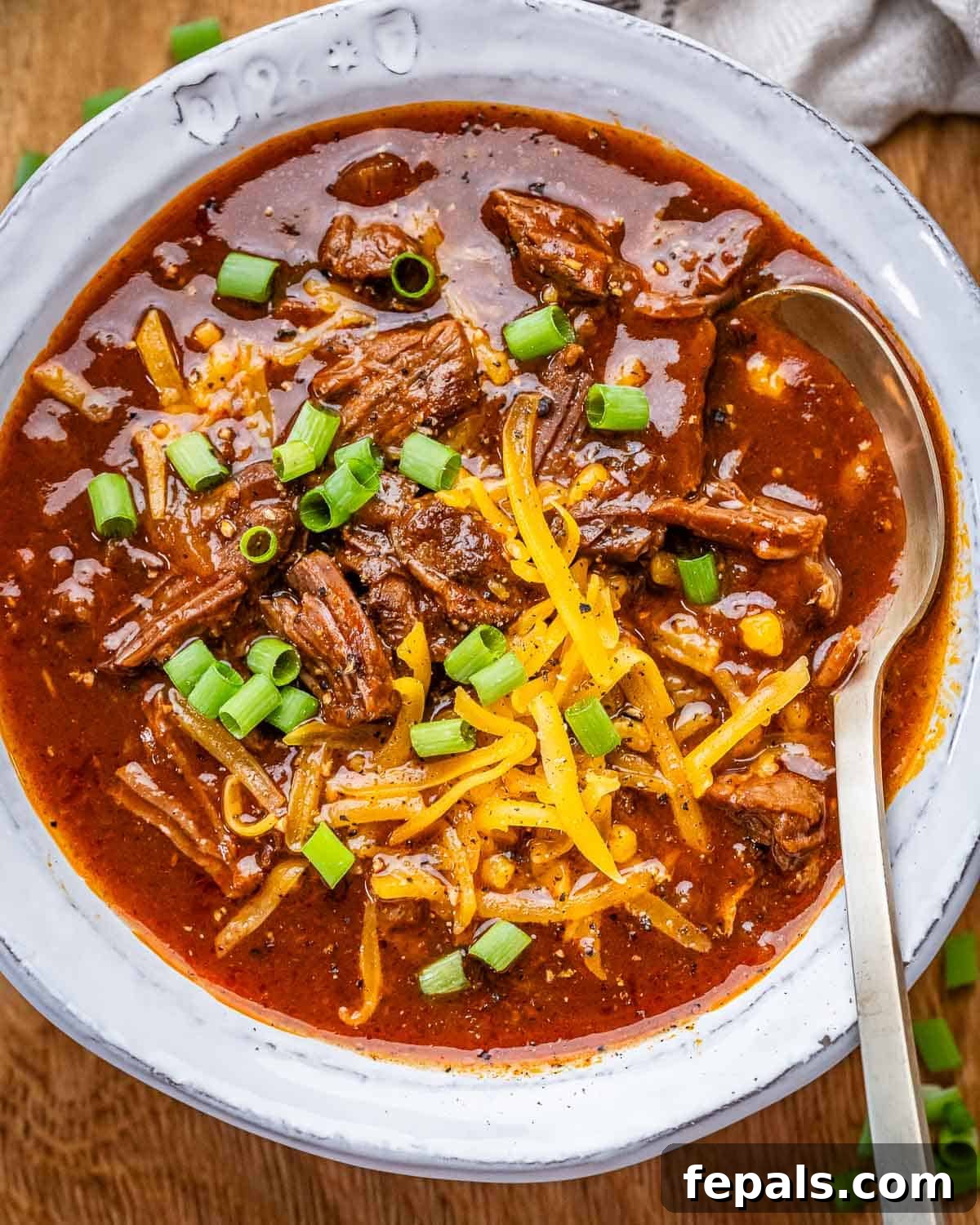 A white bowl of Texas Chili with a spoon, garnished with cheese and green onions