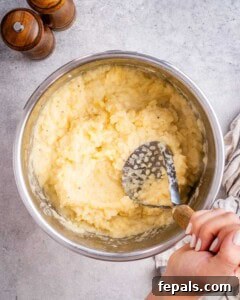Pot of freshly mashed Yukon gold potatoes being worked with a potato masher.