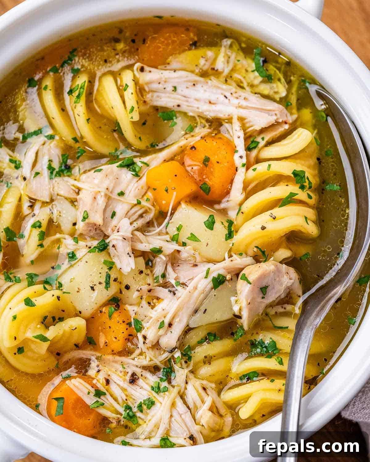 Close-up of chicken noodle soup in a white bowl, showing shredded chicken, bright orange carrots, and tender potatoes in broth.