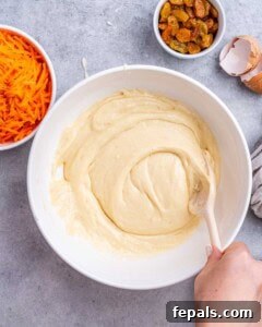 Smooth carrot cake batter in a bowl, ready to be combined with dry ingredients, with carrots and raisins in the background.