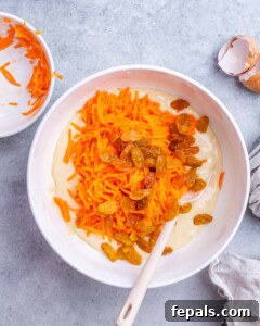 Mixing bowl with grated carrots and golden raisins being folded into batter with a spoon.