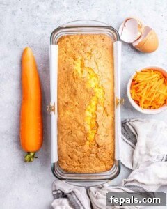 Freshly baked carrot cake loaf resting in a loaf pan, with a whole carrot and grated carrots beside it.